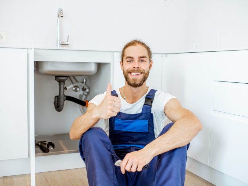 Portrait of a handsome plumber in overalls sitting near the sink at the kitchen