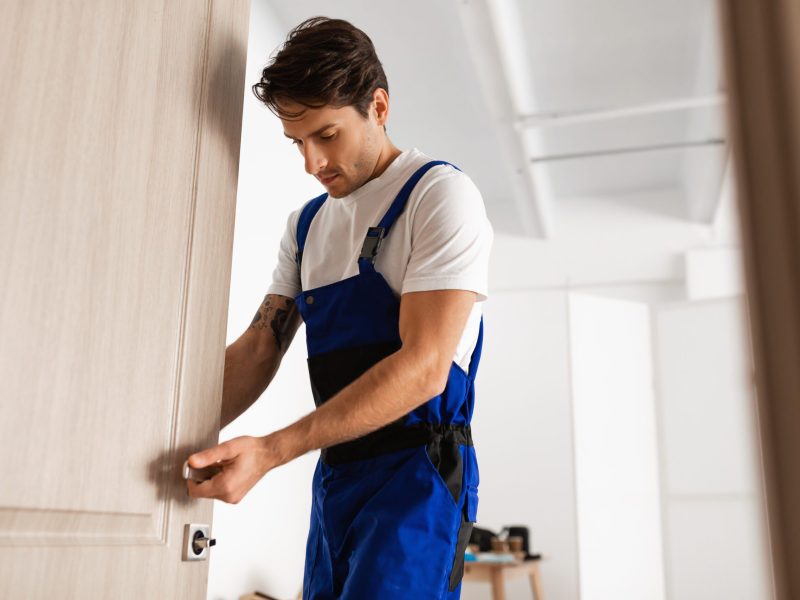 Installation of a lock on the front wooden entrance door. Portrait of young handsome locksmith workman in blue uniform installing door knob. Professional repair service. Maintenance Concept
